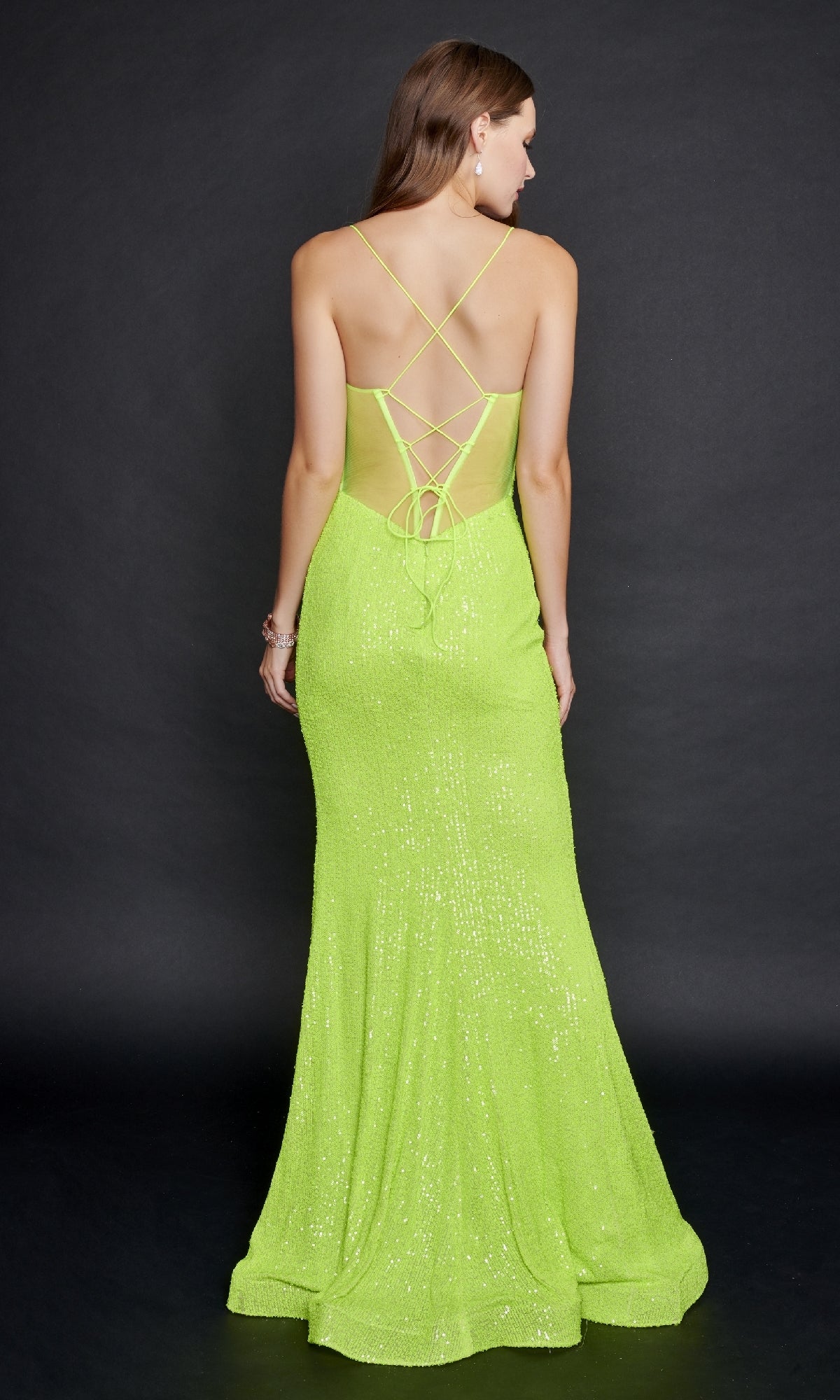 A woman faces away, wearing the Nina Canacci 8214 neon green sequin prom dress with a lace-up open back and thin straps, floor-length, against a plain dark background.